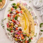 Oval platter with hummus drizzled with olive oil and sprinkled with spices, topped with mixed olives, tomatoes, feta, herbs, red onions, and served with pita bread.