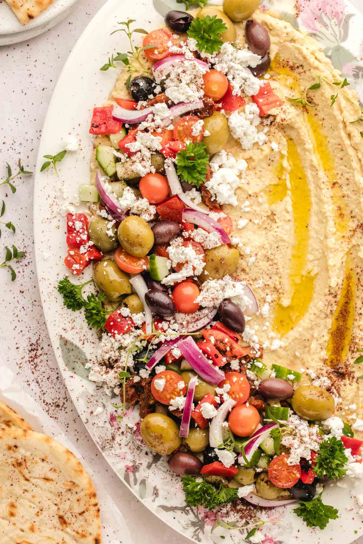 A platter of hummus topped with chopped vegetables, olives, feta cheese, and herbs, drizzled with olive oil, served with pita bread on the side.