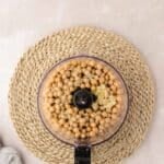 A food processor filled with chickpeas and garlic sits on a woven placemat atop a light-colored surface.