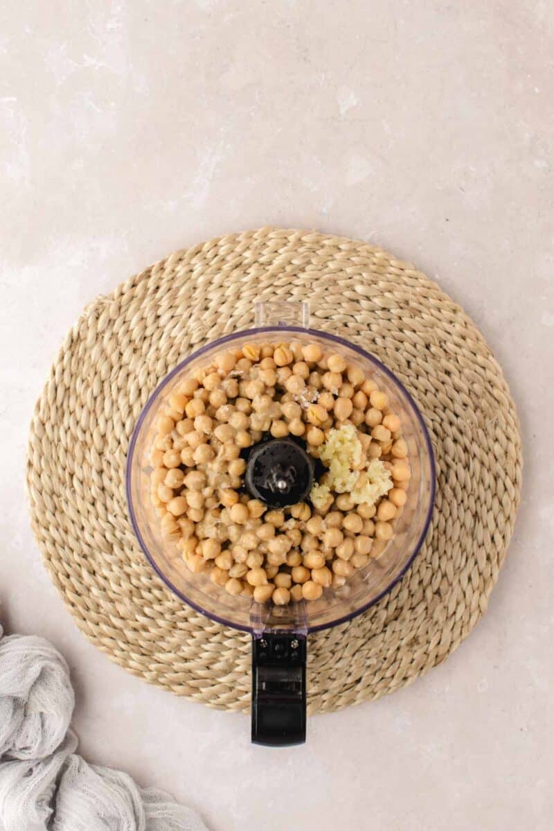 A food processor filled with chickpeas and garlic sits on a woven placemat atop a light-colored surface.