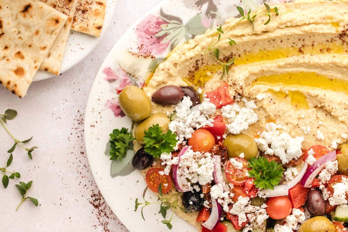A plate of hummus topped with olives, cherry tomatoes, red onion, feta cheese, parsley, and olive oil, served with pita bread on the side.
