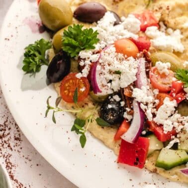 A plate with chopped tomatoes, cucumbers, red onions, olives, parsley, feta cheese, and hummus, garnished with herbs and olive oil.