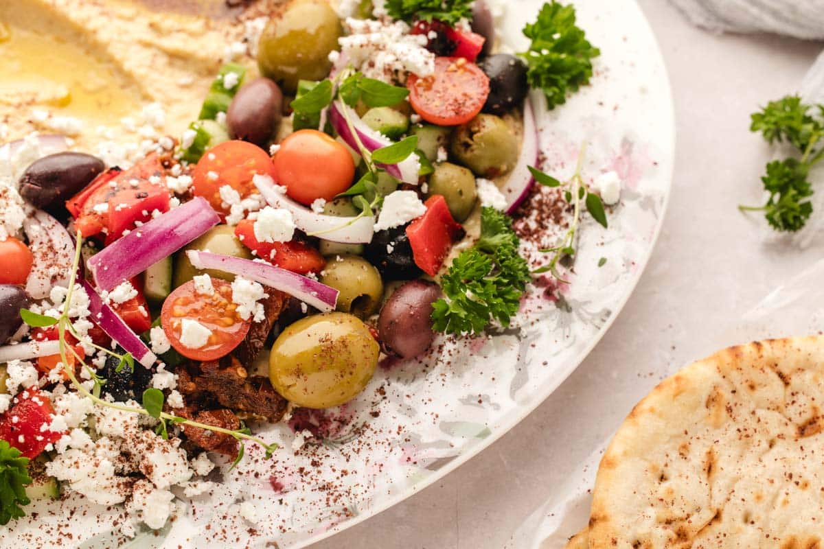 A plate with Greek salad featuring tomatoes, olives, red onions, feta cheese, parsley, and pita bread on the side.