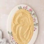 An oval plate with a floral pattern holds a smooth, spread layer of hummus, set on a light-colored surface with a light cloth nearby.