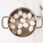A stainless steel pot with twelve white eggs arranged inside, placed on a white marble surface.