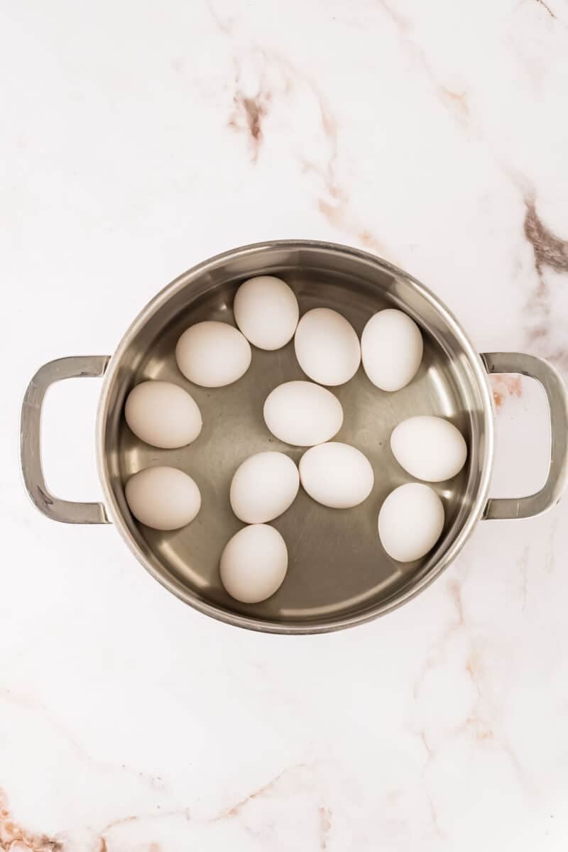 A stainless steel pot with twelve white eggs arranged inside, placed on a white marble surface.