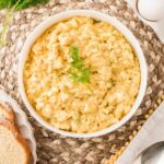 A bowl of egg salad garnished with dill sits on a woven placemat next to whole eggs, a sprig of dill, sliced bread, and a spoon.
