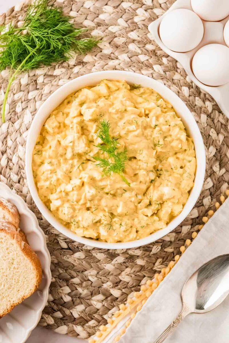 A bowl of egg salad garnished with dill sits on a woven placemat next to whole eggs, a sprig of dill, sliced bread, and a spoon.