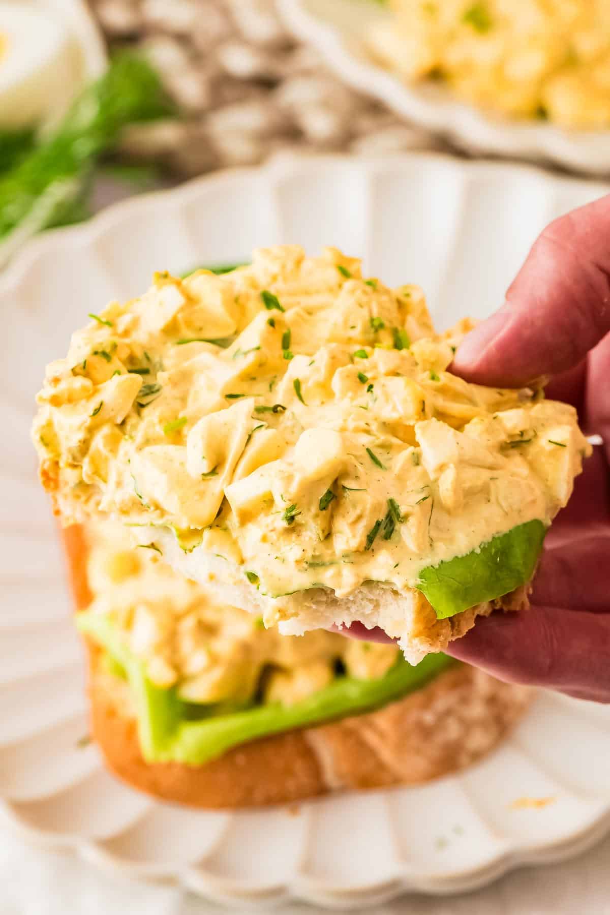 A hand holds an open-faced egg salad sandwich with sliced green bell peppers on a white plate.