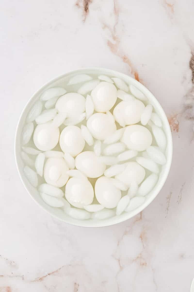 A bowl filled with peeled hard-boiled eggs and ice cubes sits on a marble surface.
