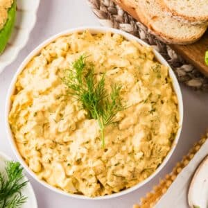 A bowl of egg salad garnished with fresh dill, surrounded by sliced bread, lettuce, and halved hard-boiled eggs.
