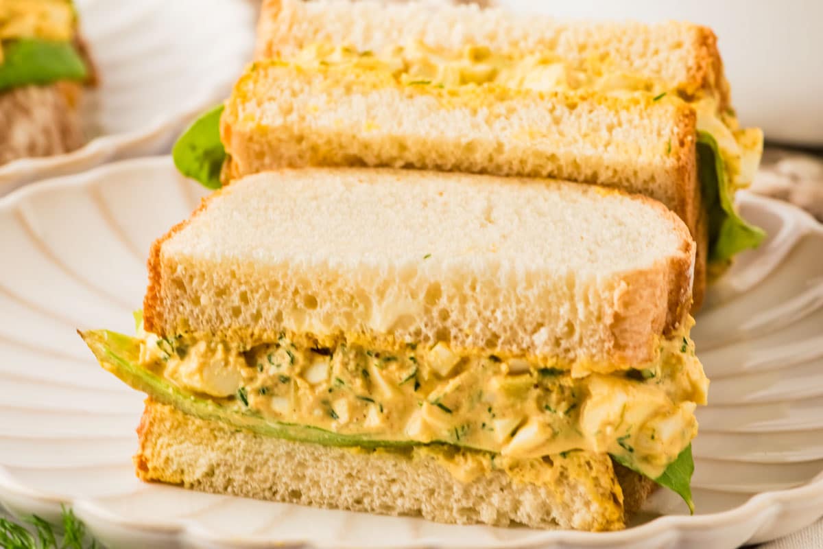 Close-up of an egg salad sandwich with leafy greens on white bread, served on a white plate.