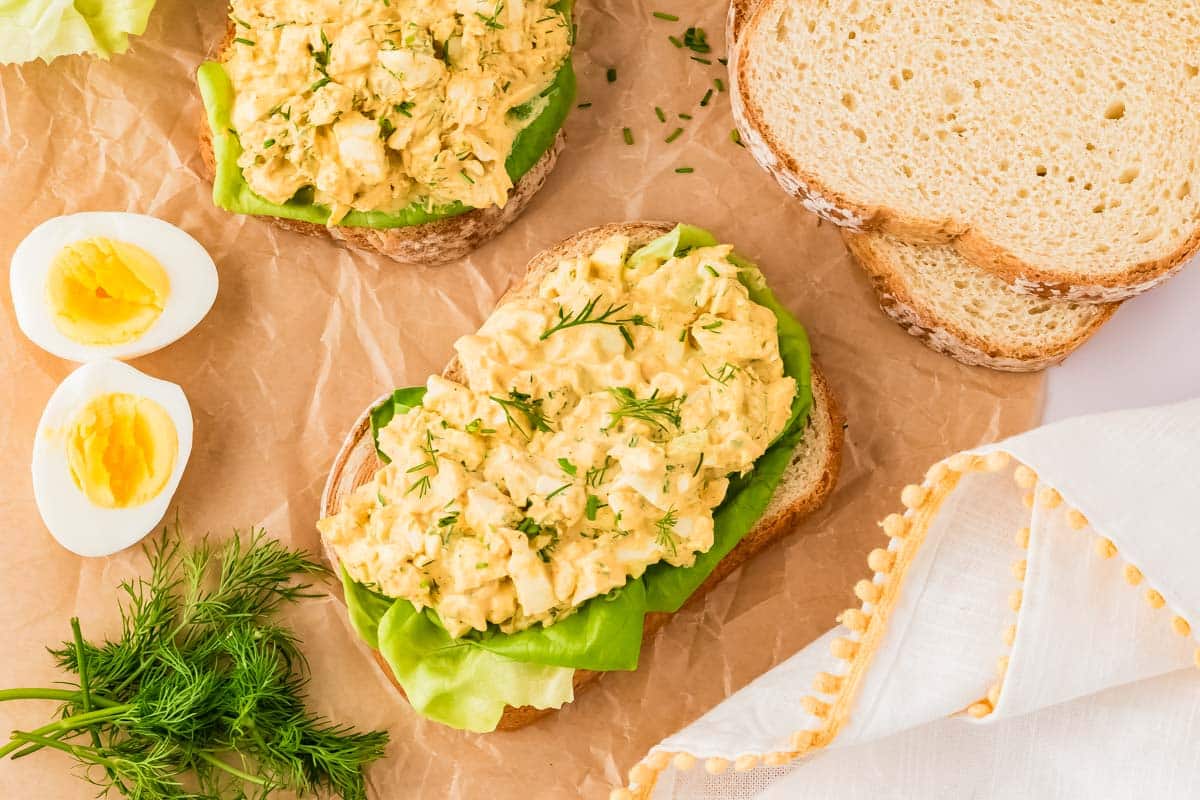 Open-faced egg salad sandwiches on slices of bread with lettuce, next to sliced hard-boiled eggs, fresh dill, and more bread on a parchment paper surface.