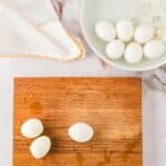 A wooden cutting board with two peeled hard-boiled eggs and one partially peeled egg, next to a bowl of water with unpeeled eggs and ice, and a white cloth.