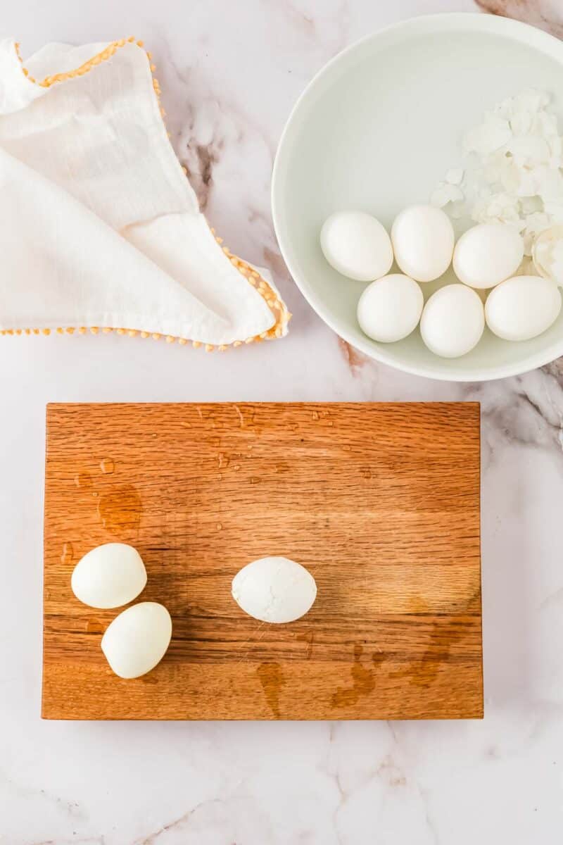 A wooden cutting board with two peeled hard-boiled eggs and one partially peeled egg, next to a bowl of water with unpeeled eggs and ice, and a white cloth.