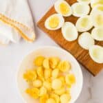Hard-boiled egg whites are on a wooden board, with egg yolks separated in a white bowl; a folded napkin lies nearby on a marble surface.