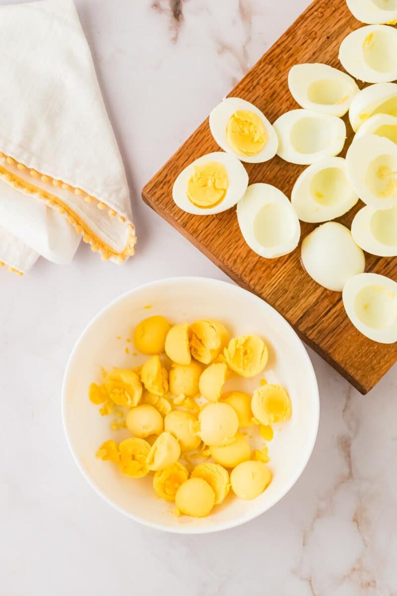 Hard-boiled egg whites are on a wooden board, with egg yolks separated in a white bowl; a folded napkin lies nearby on a marble surface.