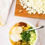 A bowl with mayonnaise, mustard, chopped celery, relish, herbs, and spices next to a cutting board with chopped hard-boiled eggs.