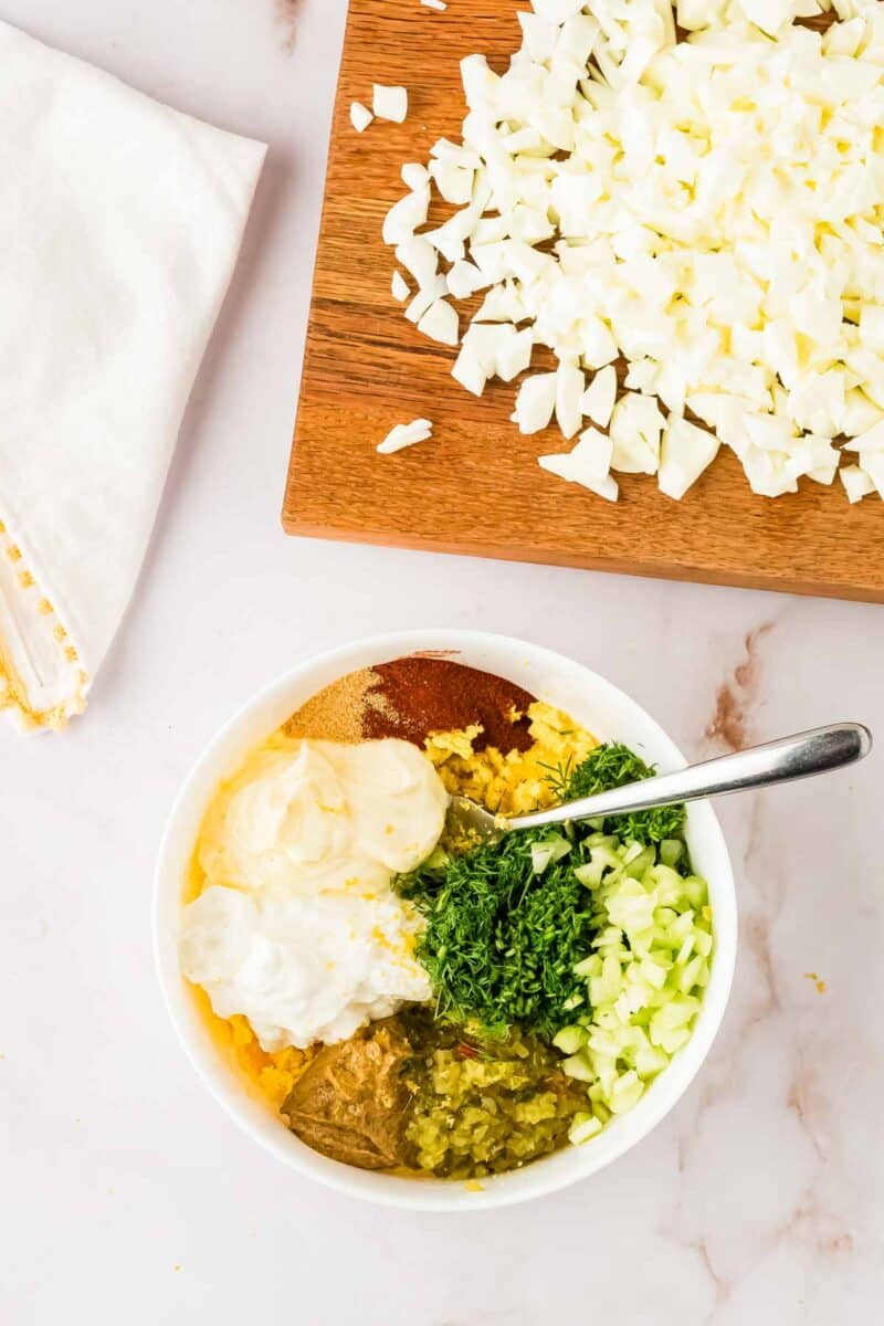 A bowl with mayonnaise, mustard, chopped celery, relish, herbs, and spices next to a cutting board with chopped hard-boiled eggs.
