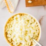 A bowl of chopped hard-boiled eggs with a spoon, next to a cutting board with more chopped eggs and a white cloth on a marble surface.
