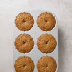 A six-cup muffin pan filled with freshly baked muffins, each with a slightly domed top and a small hole in the center, placed on a light-colored textured surface.
