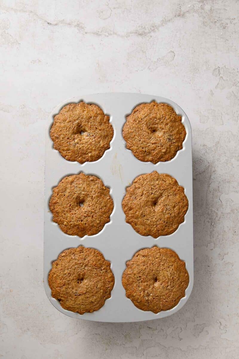 A six-cup muffin pan filled with freshly baked muffins, each with a slightly domed top and a small hole in the center, placed on a light-colored textured surface.