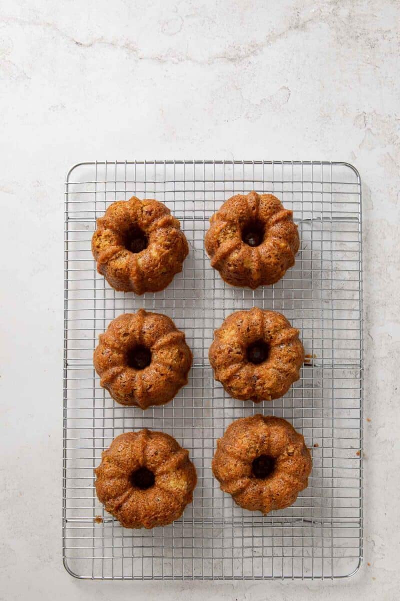 Six mini Bundt cakes are arranged in two rows on a wire cooling rack set on a light-colored surface.