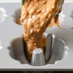 Thick batter is being poured from a spatula into a greased, metal mini Bundt cake pan cavity.