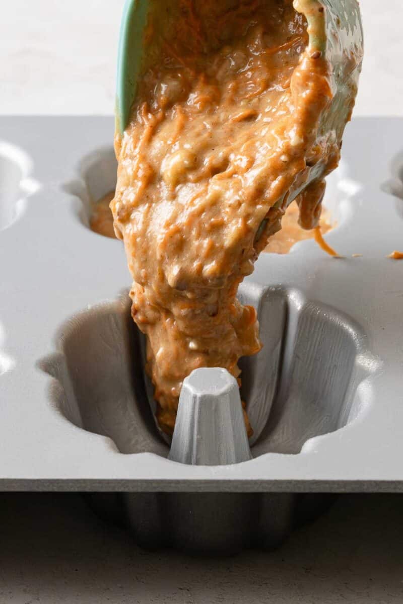Thick batter is being poured from a spatula into a greased, metal mini Bundt cake pan cavity.
