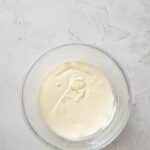 A glass bowl containing a thick, pale cream mixture sits on a light textured surface.