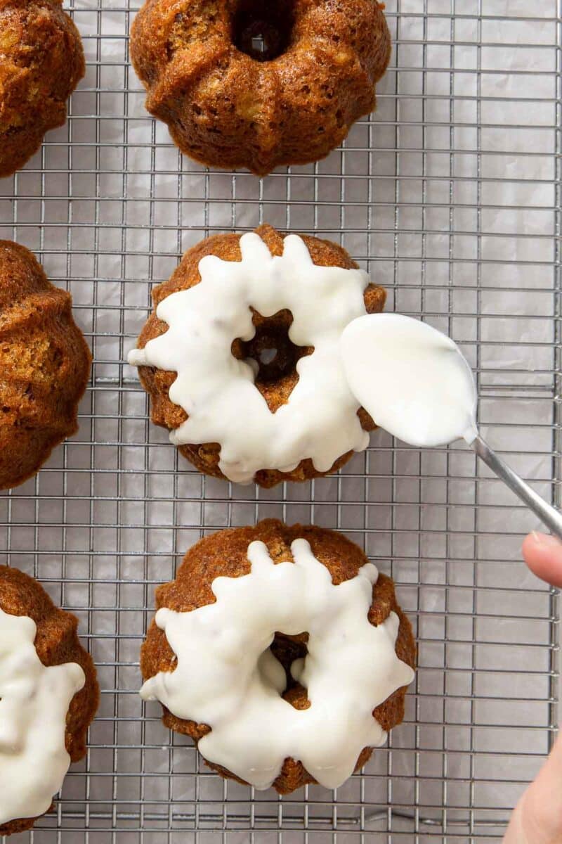 A hand uses a spoon to drizzle white icing onto mini bundt cakes arranged on a wire cooling rack.