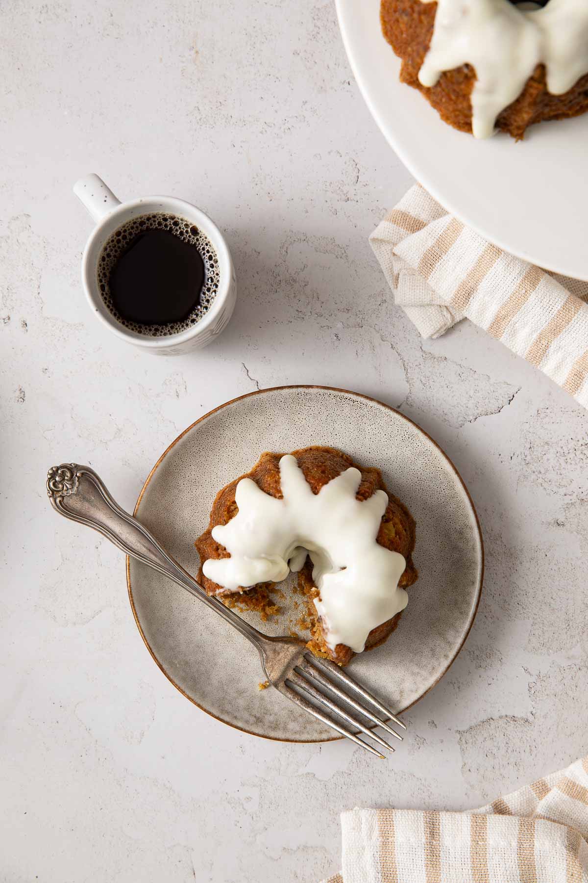 A small bundt cake with white icing on a plate with a fork, next to a cup of black coffee and a striped napkin on a white surface.