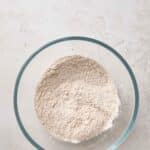 A glass bowl filled with a mixture of dry flour on a light-colored textured surface.