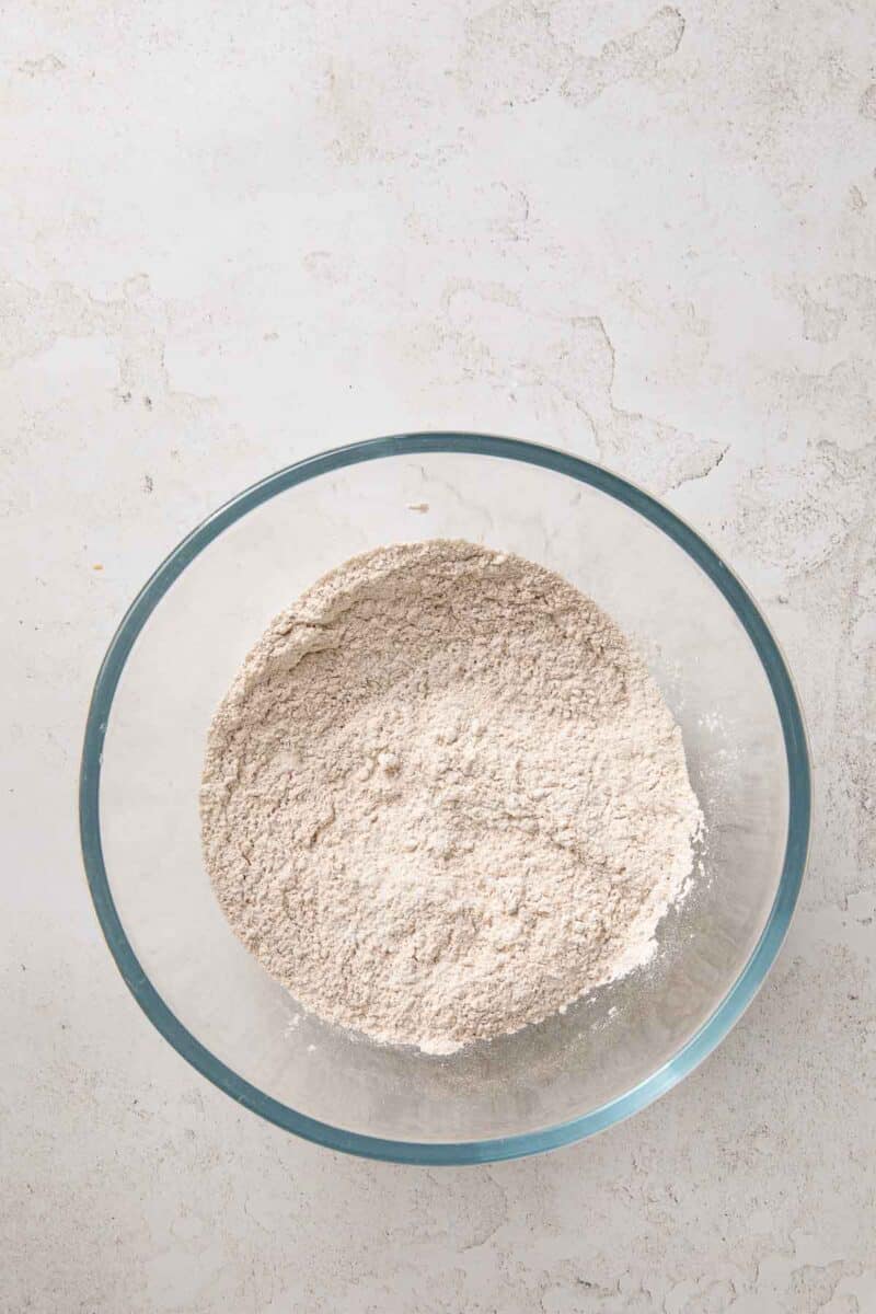 A glass bowl filled with a mixture of dry flour on a light-colored textured surface.