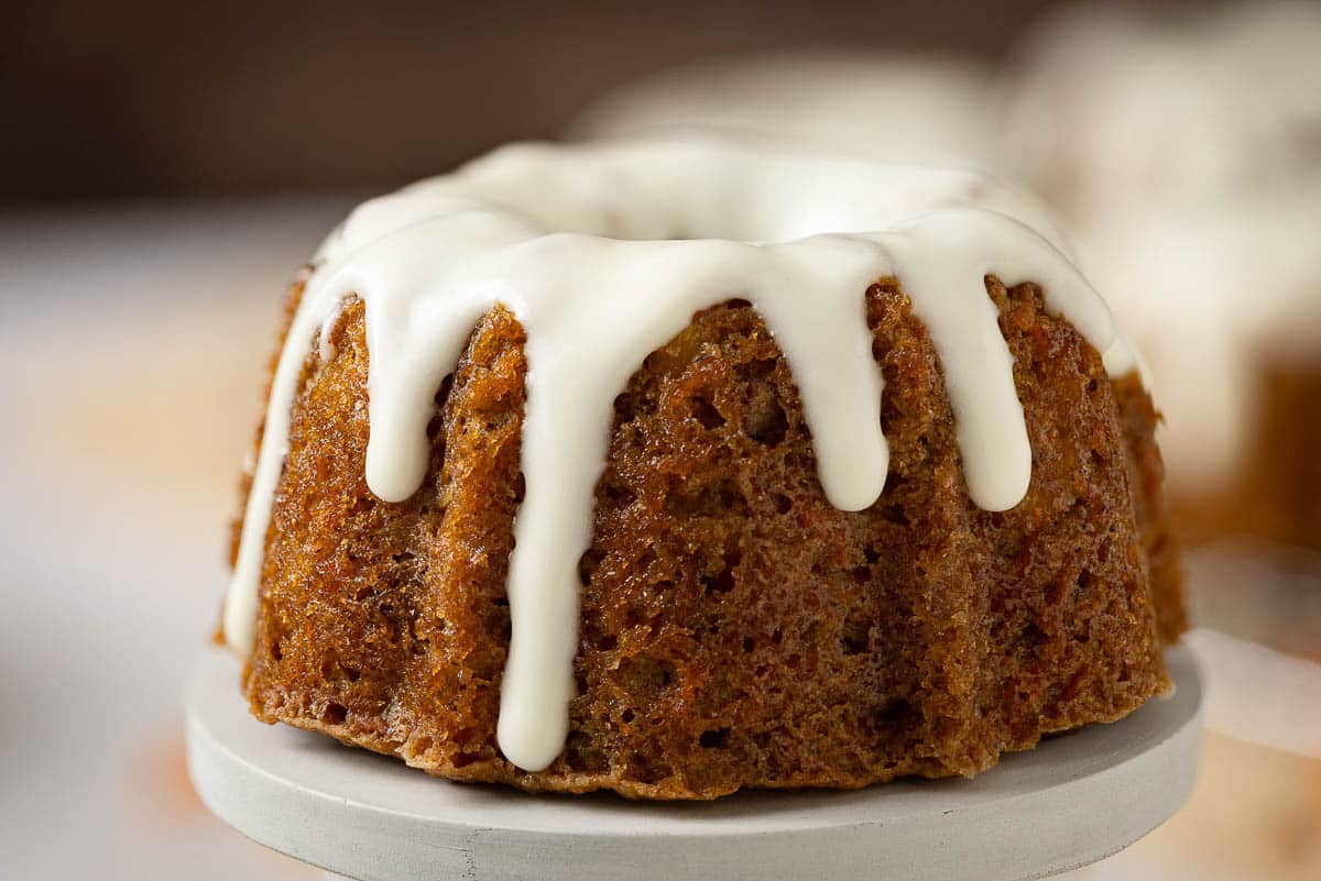 A small bundt cake with white icing drizzled on top, displayed on a white stand.