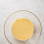 A clear glass bowl containing a light yellow batter sits on a textured white surface.