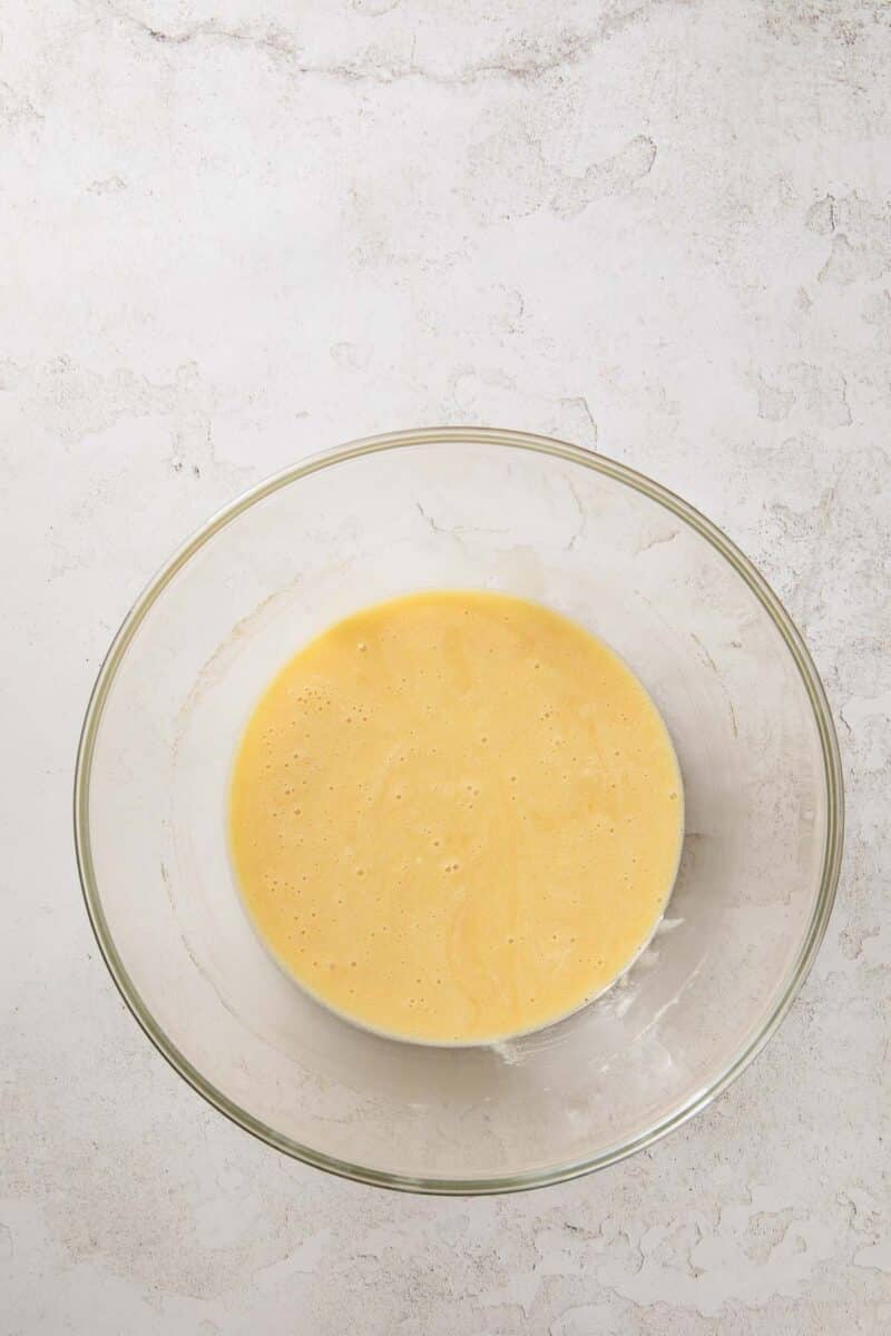 A clear glass bowl containing a light yellow batter sits on a textured white surface.