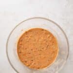 A glass mixing bowl filled with a light brown batter containing visible shredded carrots and small bits, placed on a white textured surface.