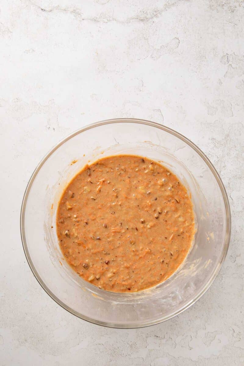 A glass mixing bowl filled with a light brown batter containing visible shredded carrots and small bits, placed on a white textured surface.