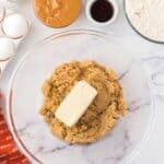 A glass mixing bowl with brown sugar and a stick of butter, surrounded by eggs, peanut butter, milk, vanilla, flour, and peanut butter cups on a white countertop.