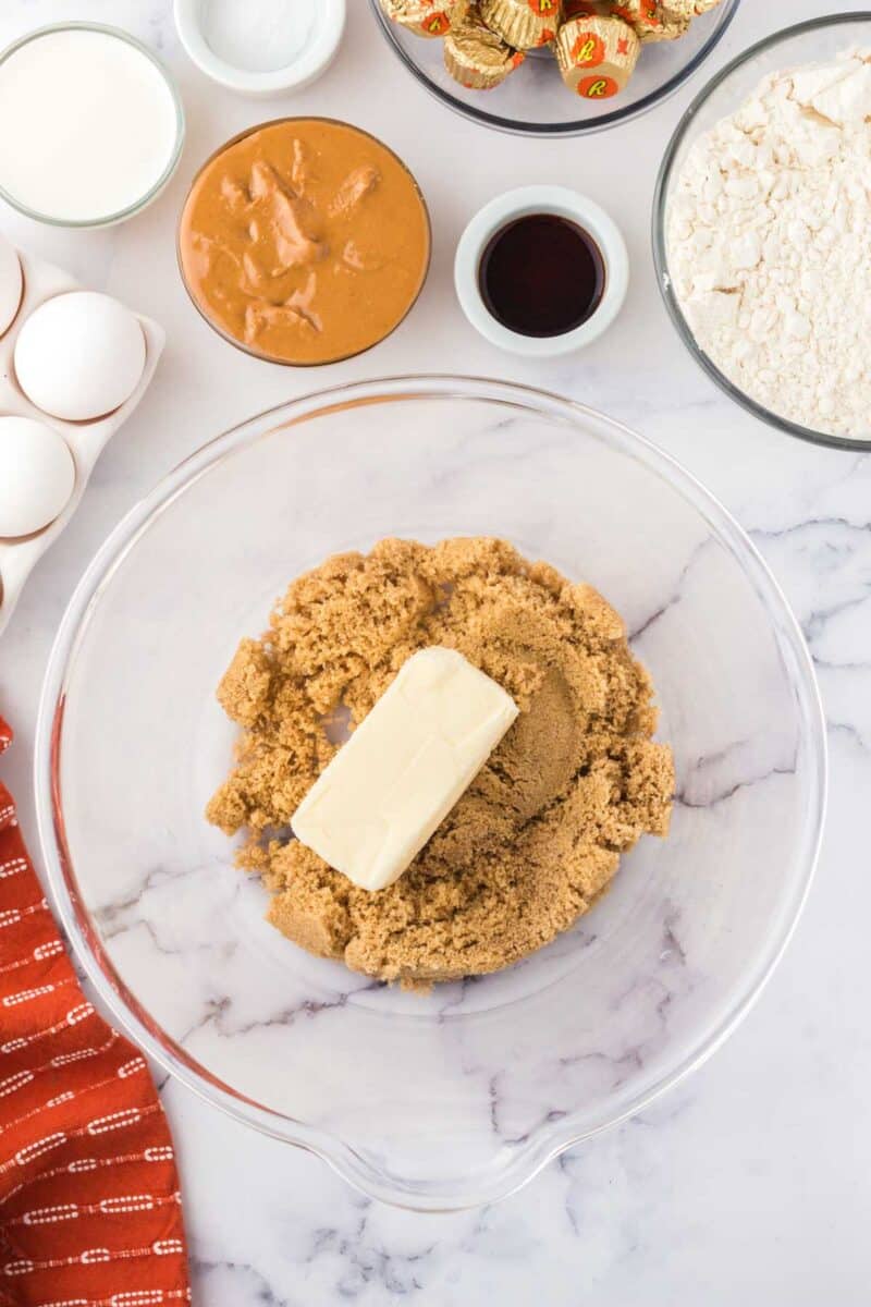 A glass mixing bowl with brown sugar and a stick of butter, surrounded by eggs, peanut butter, milk, vanilla, flour, and peanut butter cups on a white countertop.