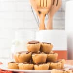 A plate of peanut butter cup cookies is stacked on a kitchen counter, with wooden utensils and jars in the background.