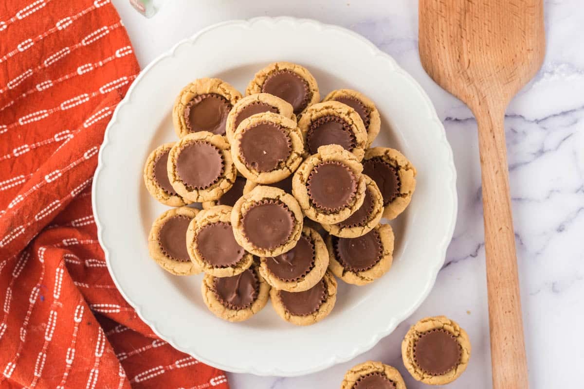 A white plate holds a pile of peanut butter cup cookies on a marble surface, next to a wooden spatula and an orange patterned cloth.