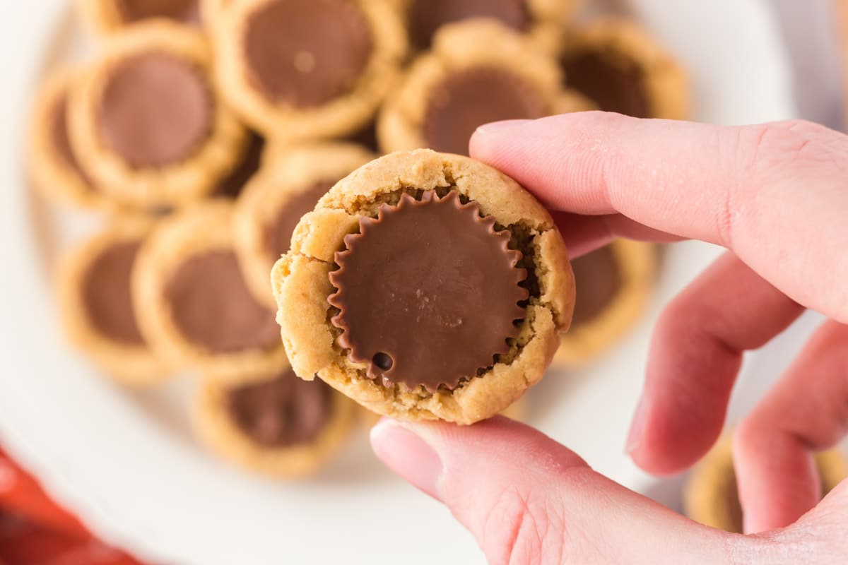 A hand holding a peanut butter cookie with a chocolate peanut butter cup center, with more cookies on a plate in the background.