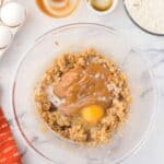 A mixing bowl contains partially mixed cookie dough with brown sugar, eggs, peanut butter, and milk, surrounded by baking ingredients on a marble countertop.