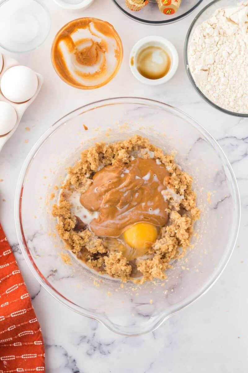 A mixing bowl contains partially mixed cookie dough with brown sugar, eggs, peanut butter, and milk, surrounded by baking ingredients on a marble countertop.