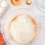 A glass mixing bowl with flour on top of peanut butter mixture, surrounded by eggs, vanilla extract, peanut butter, and candy cups on a marble countertop.