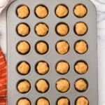 A muffin tin filled with raw dough balls sits on a marble countertop, surrounded by eggs, empty bowls, and an orange kitchen towel.