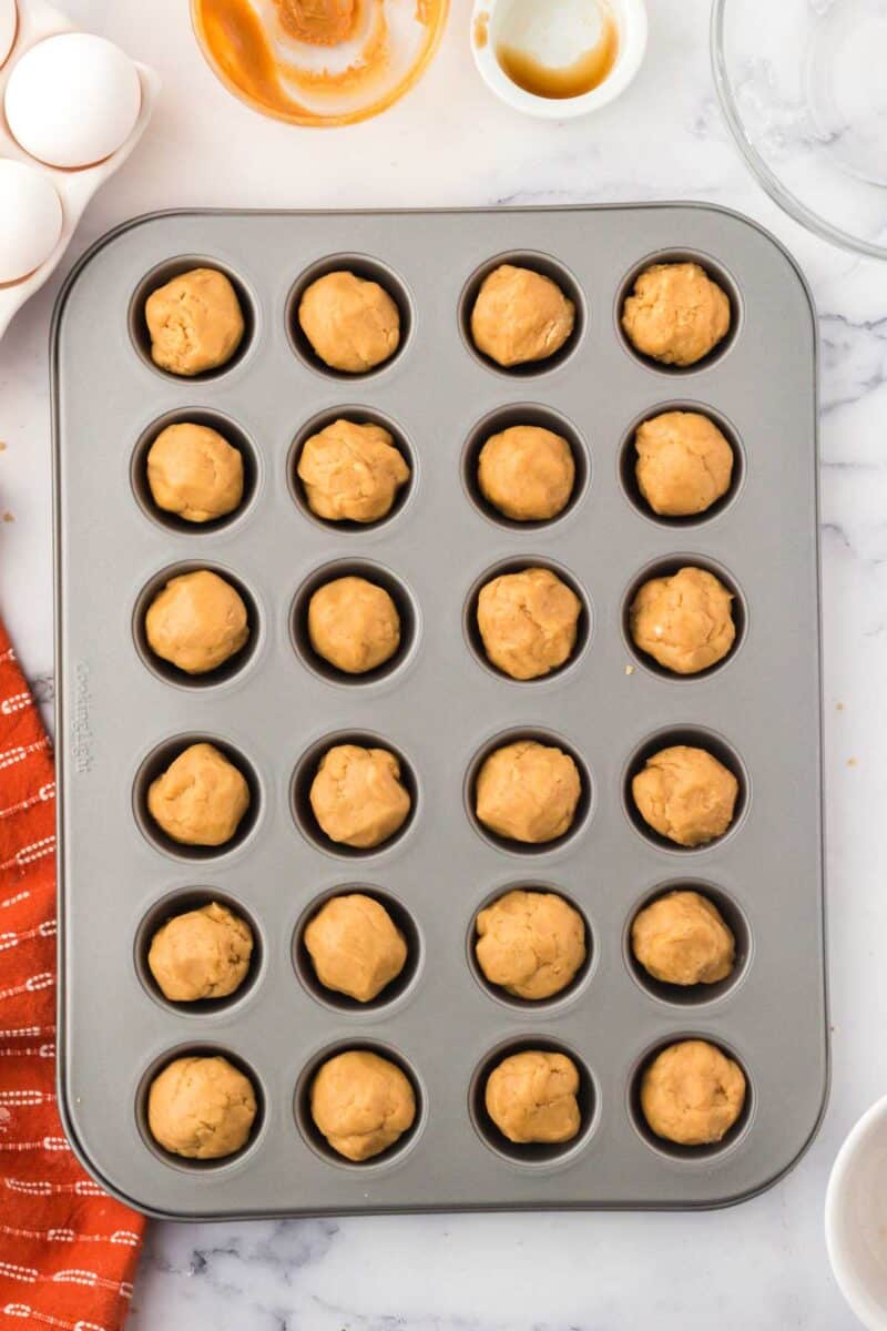 A muffin tin filled with raw dough balls sits on a marble countertop, surrounded by eggs, empty bowls, and an orange kitchen towel.