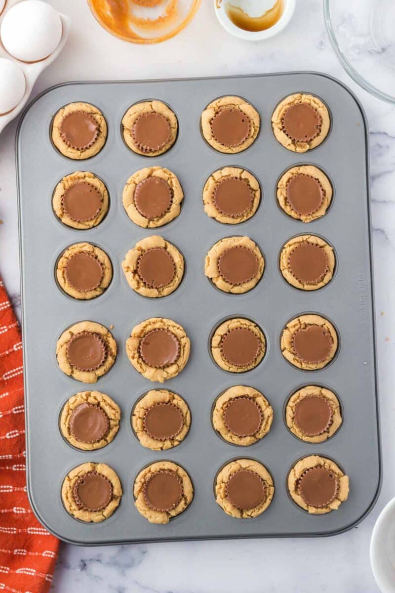 A muffin tin filled with 24 peanut butter cup cookies, each topped with a chocolate peanut butter cup, on a marble countertop.